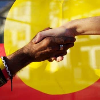 Black and white people shaking hands in front of the Aboriginal Flag