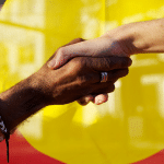 Black and white hands joined in front of the Aboriginal flag